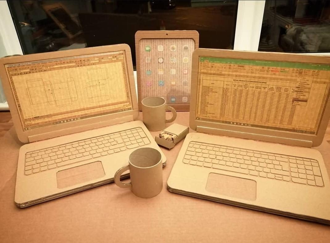 Cardboard replicas of laptops, a tablet, and a mug arranged on a table.